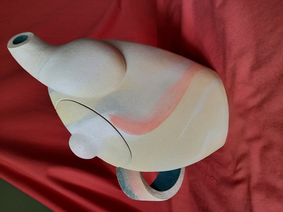 Artistic ceramic teapot with a pastel blend of beige, pink, and light blue, featuring a wide spout and arched handle, displayed against a red fabric backdrop.